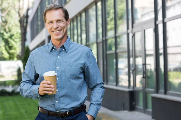 Sonriente hombre de negocios posando con taza de café desechable cerca del edificio de oficinas - foto de stock
