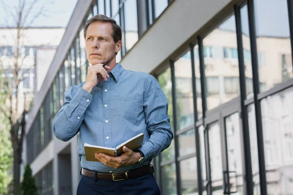 Homem de negócios pensativo com diário e caneta de pé perto do prédio de escritórios — Fotografia de Stock