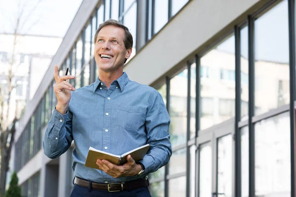 Homme d'affaires souriant avec journal ayant une idée et pointant vers le haut près de l'immeuble de bureaux — Photo de stock