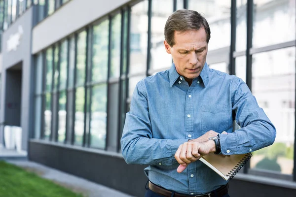 Mature businessman with diary looking at wristwatch near office building — Stock Photo
