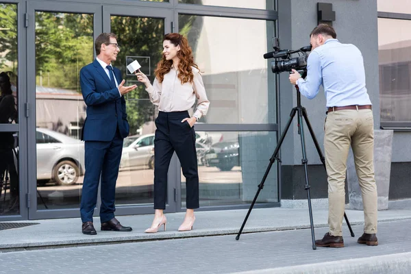 Professional cameraman and news reporter interviewing businessman near office building — Stock Photo