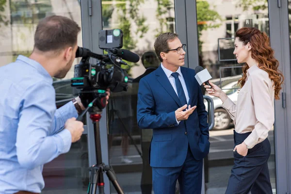 Cameraman and news reporter with microphone interviewing businessman near office building — Stock Photo