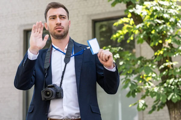 Reportero gráfico masculino con cámara fotográfica digital haciendo gestos y mostrando pase de prensa - foto de stock