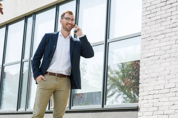 Smiling businessman in formal wear talking on smartphone near office building — Stock Photo