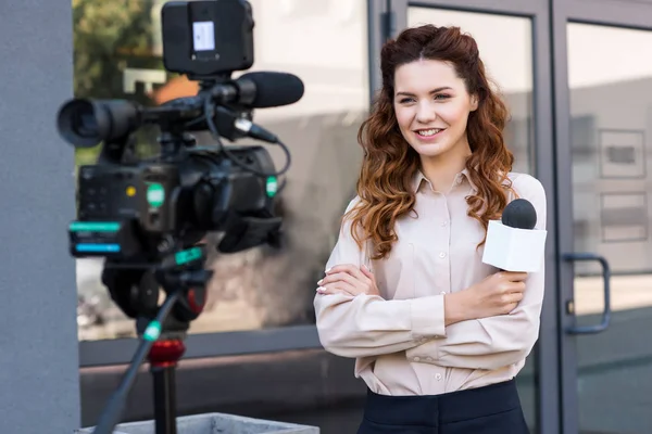 Giornalista sorridente con microfono in piedi davanti alla videocamera digitale — Foto stock