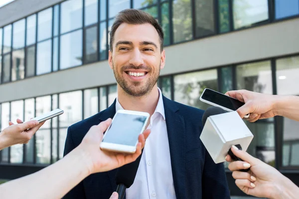 Journalists interviewing smiling successful businessman with microphones and smartphones — Stock Photo