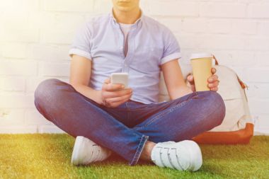 gitmek için smartphone ve kahve ile çimenlerin üzerinde oturan genç adam resmini kırpılmış
