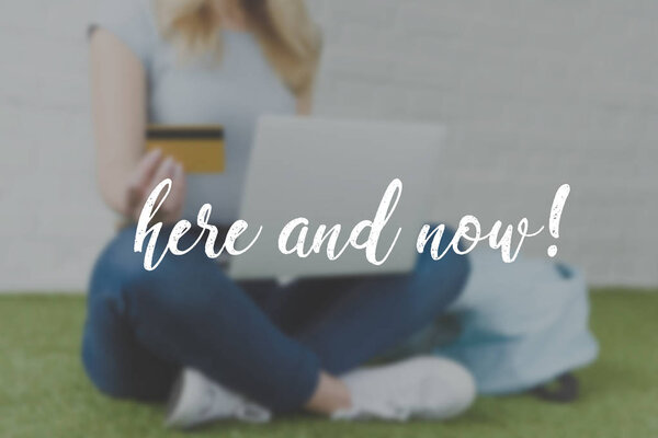 обрезанный снимок молодой женщины, делающей электронные покупки с ноутбуком, сидя на траве, здесь и сейчас надпись
