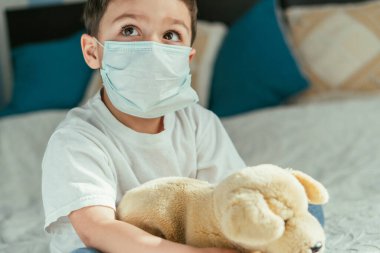 Tıbbi maskeli şirin çocuk evde yumuşak oyuncaklar tutuyor.