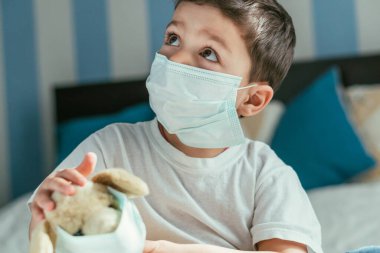Tıbbi maskeli küçük bir çocuğun elinde yumuşak bir oyuncakla yukarı baktığı seçici odaklanma.