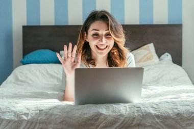 Neşeli bir kadın yatak odasında video görüşmesi yaparken el sallıyor. 