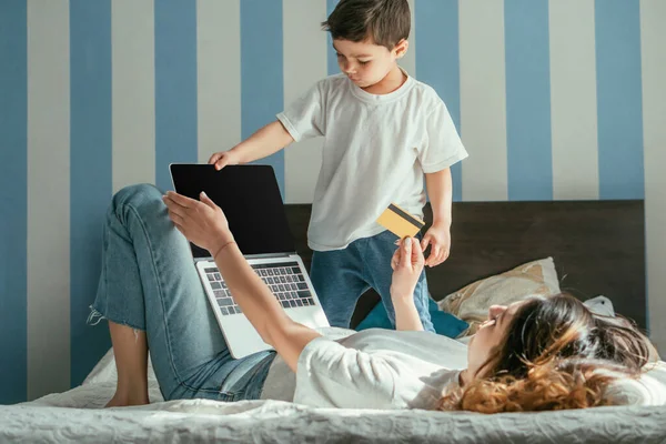 女性躺在床上 带着空白屏幕和蹒跚学步的男婴在笔记本电脑旁拿着信用卡的选择性焦点 — 图库照片