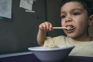 Tatlı esmer çocuk mutfak masasının yanında çatallı makarna yiyor.