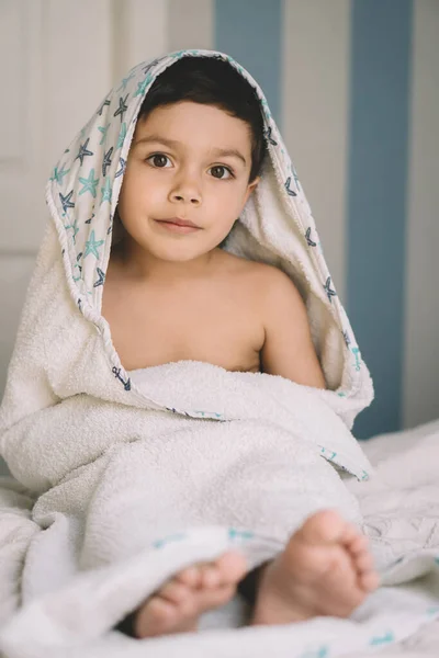 かわいい笑顔の男の子の選択的な焦点フードタオルに包まれベッドに座っている間カメラを見て — ストック写真