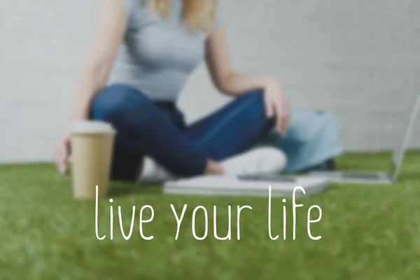 Cropped shot of woman sitting on grass with cup of coffee to go and working with laptop, live your life inscription — Stock Photo