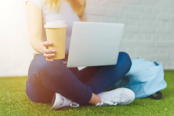 Tiro recortado de jovem mulher sentada na grama com xícara de café para ir e trabalhar com laptop — Fotografia de Stock