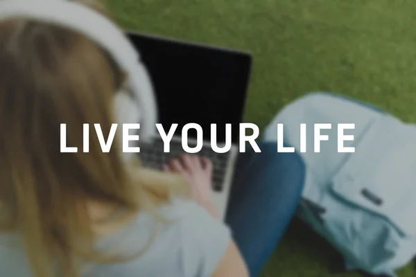 High angle view of young woman sitting on grass with headphones on head and working with laptop, live your life inscription — Stock Photo