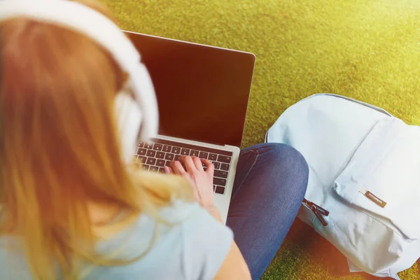 Vue grand angle de la jeune femme assise sur l'herbe avec écouteurs sur la tête et de travailler avec ordinateur portable — Photo de stock