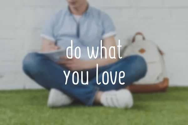 Cropped shot of young man writing in notebook while sitting on grass with backpack, do what you love inscription — Stock Photo