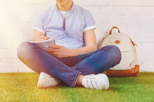 Recortado disparo de hombre joven escribiendo en el cuaderno mientras está sentado en la hierba con la mochila - foto de stock