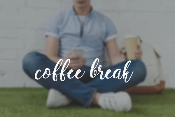 Tiro cortado de jovem sentado na grama com smartphone e café para ir, inscrição coffee break — Fotografia de Stock