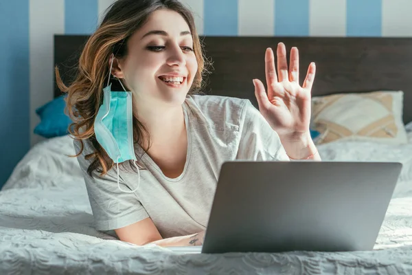 Mujer alegre en máscara médica saludando la mano mientras tiene videollamada en casa — Stock Photo