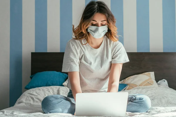 Freelancer en máscara médica usando laptop en la cama - foto de stock