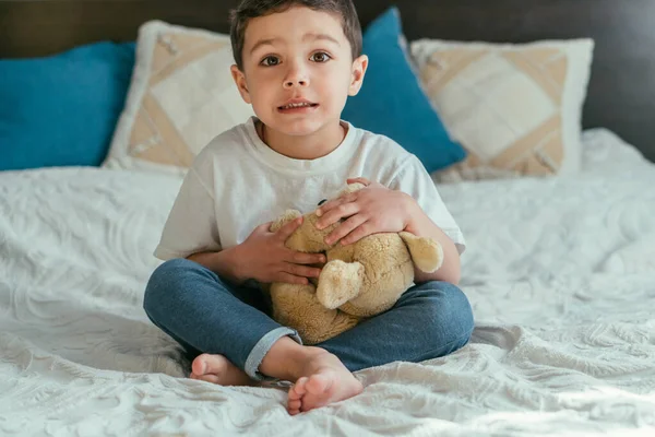 Cute toddler boy with barefoot sitting on bed with soft toy — Stock Photo