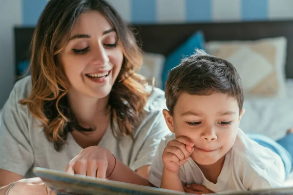 Selektiver Fokus eines Kleinkindes, das nahe der Mutter lächelt, die zu Hause Buch liest — Stockfoto