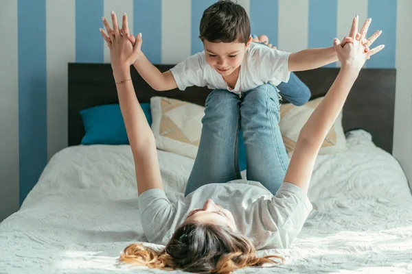 Foco seletivo da mãe deitada na cama e de mãos dadas com o filho criança alegre enquanto joga no quarto — Fotografia de Stock