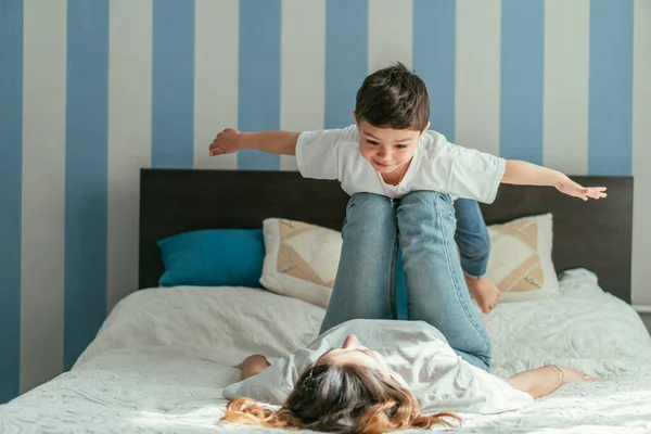 Selective focus of toddler son with outstretched hands lying on legs of mother in bedroom — Stock Photo