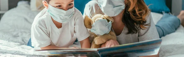 Panoramic concept of mother and toddler son in medical masks reading book near soft toy — Stock Photo