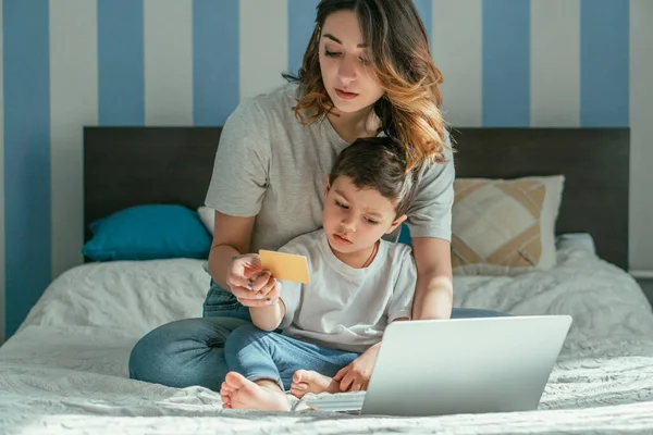 Mulher segurando cartão de crédito enquanto sentado na cama com o filho da criança perto do laptop — Fotografia de Stock
