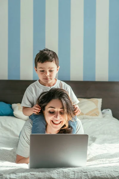 Mignon tout-petit garçon toucher les cheveux de joyeux pigiste mère en utilisant un ordinateur portable dans la chambre — Photo de stock