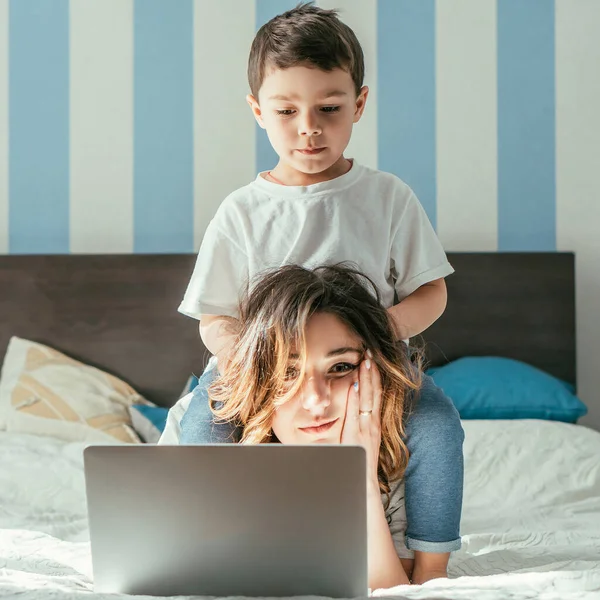 Lindo niño tocando el pelo de la madre freelancer cansado cerca del ordenador portátil en el dormitorio - foto de stock