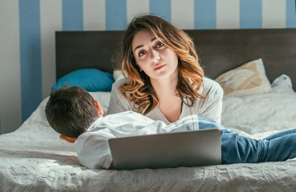 Menino criança deitada na cama perto de mãe freelancer cansado e laptop — Fotografia de Stock