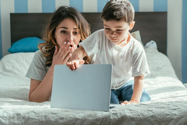 Sorprendido freelancer cubriendo la boca y mirando a su hijo pequeño tocando portátil en el dormitorio - foto de stock