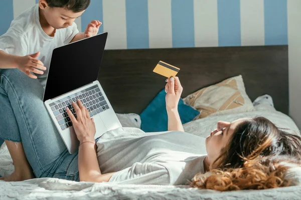 Foco seletivo da mulher deitada na cama e segurando cartão de crédito perto de laptop com tela em branco e filho criança — Fotografia de Stock