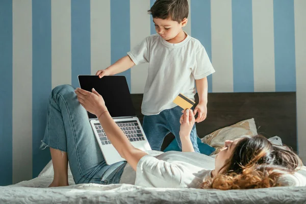 Selective focus of woman lying on bed and holding credit card near laptop with blank screen and toddler boy — Stock Photo