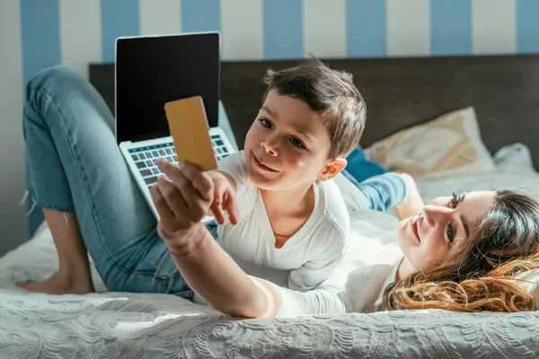 Selektiver Fokus der Frau, die auf dem Bett liegt und Kreditkarte in der Nähe von Laptop mit leerem Bildschirm und niedlichem Kleinkind hält — Stockfoto