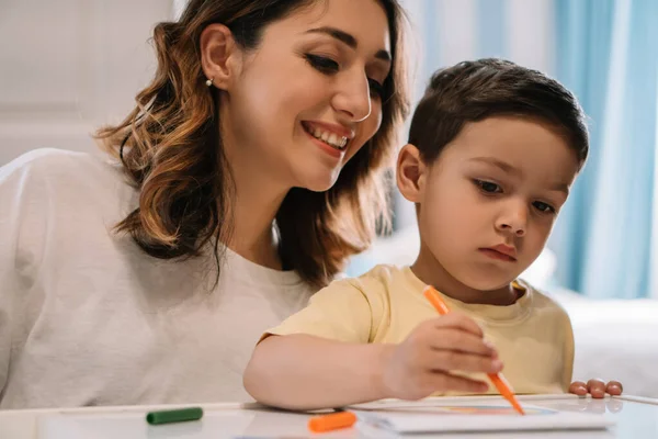 Cheerful mother looking at adorable son drawing with felt-tip pen — Stock Photo