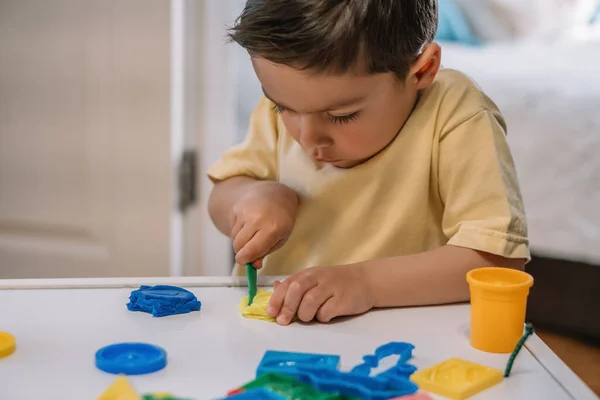 Aufmerksamer, entzückender Junge schneidet bunte Knetmasse mit Spachtel — Stockfoto