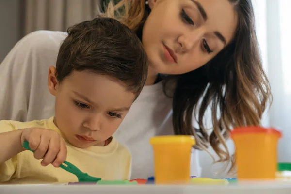 Junge Mutter sieht konzentrierten Sohn mit Spachtel in der Nähe von Knetmasse — Stockfoto