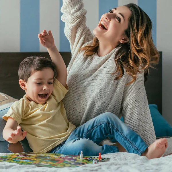 Feliz, madre emocionada e hijo mostrando gestos ganadores mientras juega juego de mesa en la cama - foto de stock