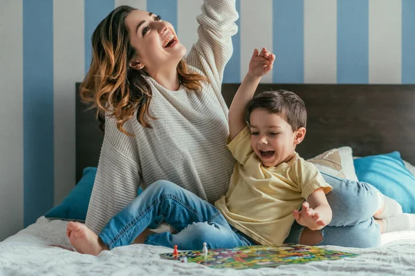 Emocionados madre e hijo mostrando gestos ganadores mientras juega juego de mesa en la cama - foto de stock