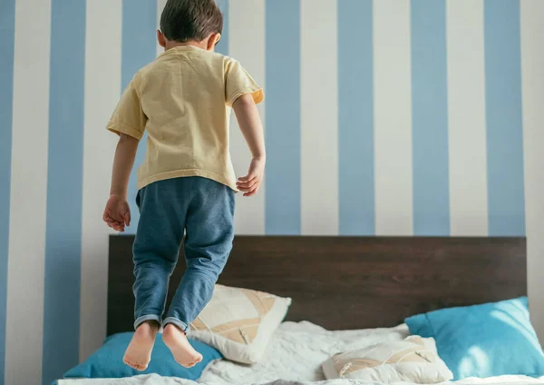 Back view of barefoot boy in blue jeans jumping on bed at home — Stock Photo