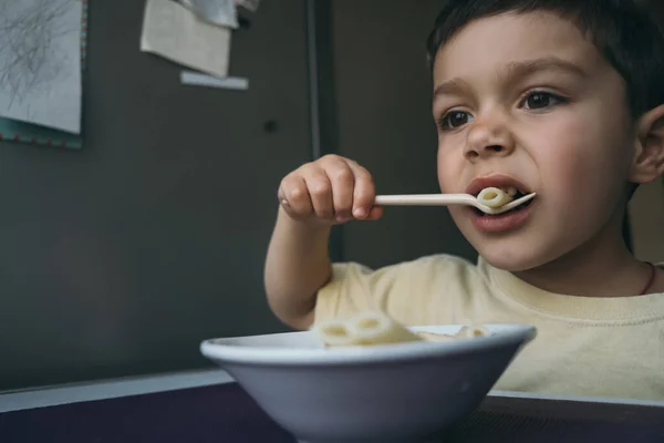 Милый брюнетка мальчик ест макароны с вилкой возле миски на кухонном столе — стоковое фото