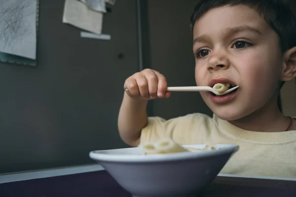 Niedliche brünette junge essen penne pasta mit gabel in der nähe von schüssel auf küchentisch — Stockfoto