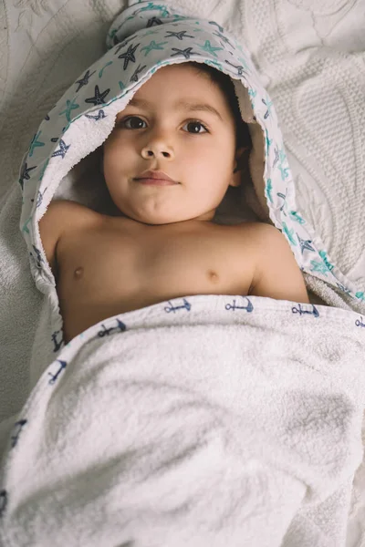 Vista superior de un niño adorable, envuelto en una toalla con capucha, acostado en la cama y mirando a la cámara - foto de stock