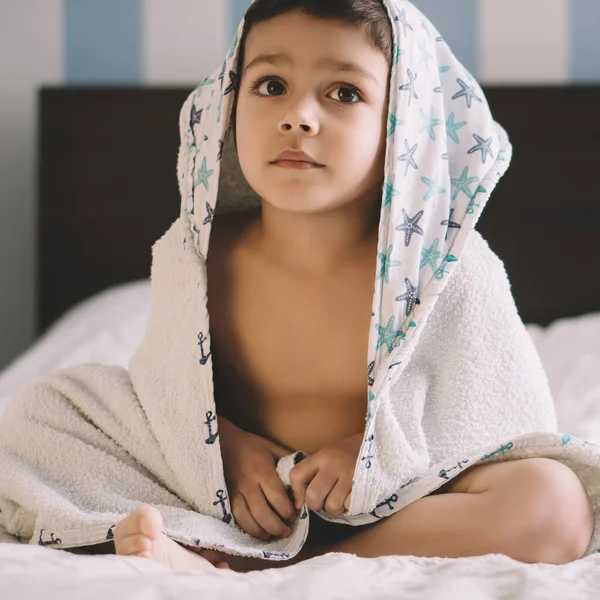 Menino adorável, coberto com toalha com capuz, sentado na cama e olhando para longe — Fotografia de Stock